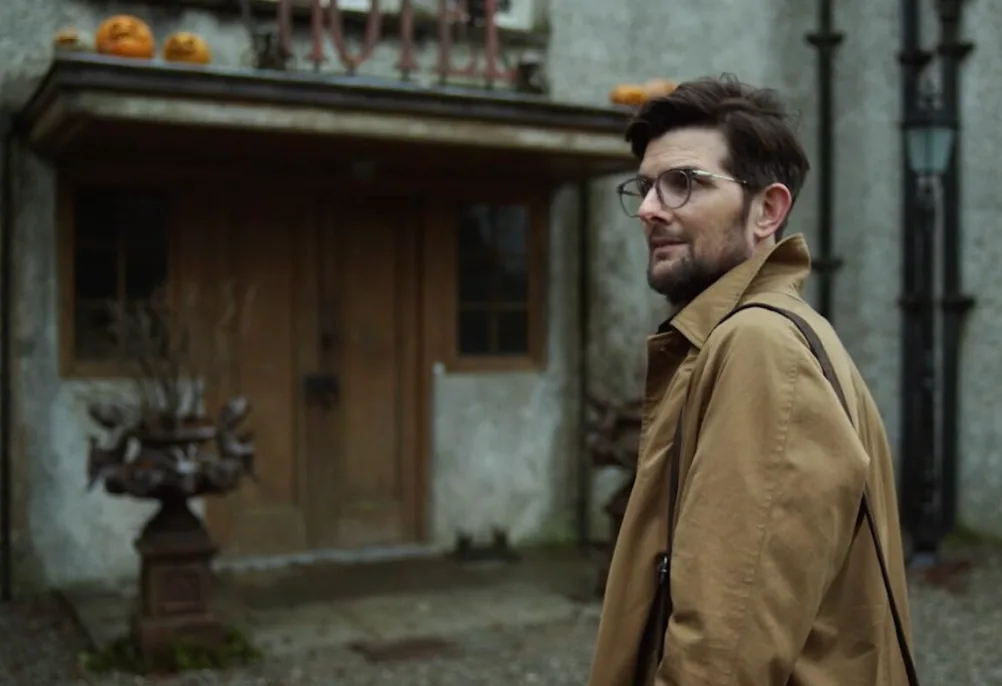 A man wearing glasses and a tan coat outside an old stone building in Hokum.