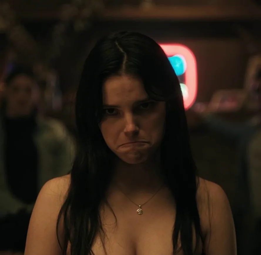 A dark-haired woman frowning in a crowded room with neon lighting in Obsession.