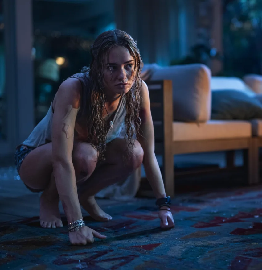 A woman crouching on a patterned rug in a dimly lit room in Primate.