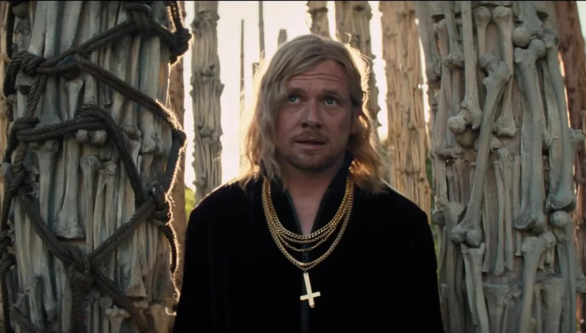 A blond man wearing gold chains and a cross stands between pillars made of bones in 28 Years Later: The Bone Temple.