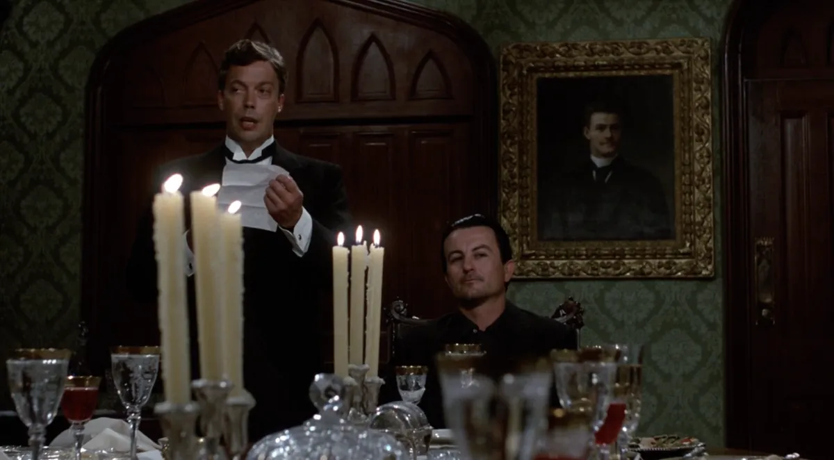 Tim Curry reads a note beside a candlelit dining table in Clue (1985).
