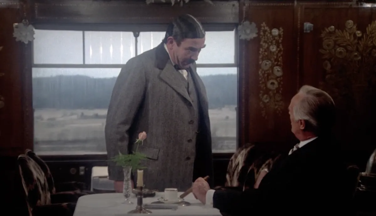 Albert Finney as Poirot beside a seated passenger in a train dining car in Murder on the Orient Express (1974).