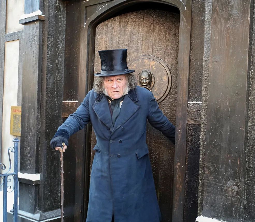 A man in a blue coat and top hat holds a cane in a wooden doorway.