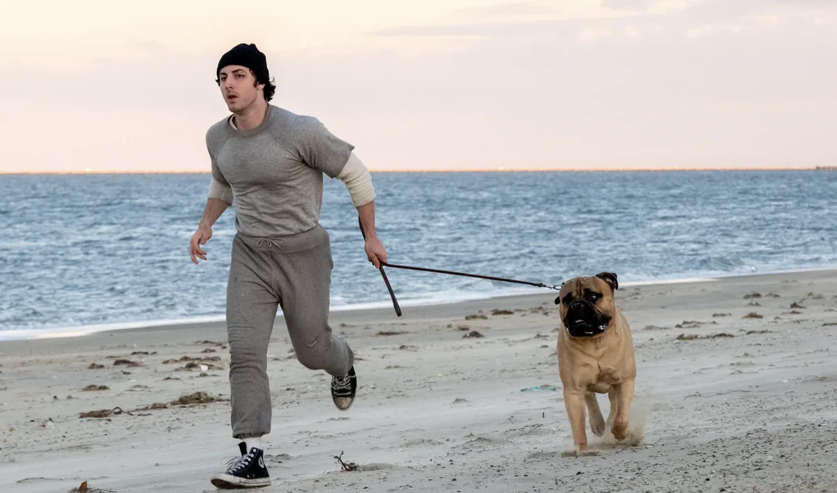 A young man in gray sweats jogs along a beach with a dog on a leash.