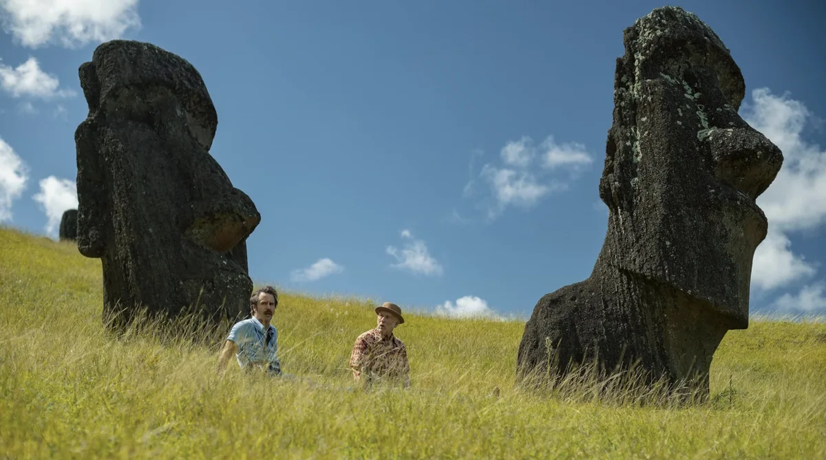 Two men stand in tall grass between large moai statues on Easter Island.