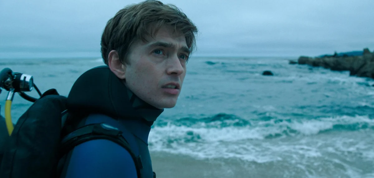 A diver wearing a blue wetsuit and scuba gear beside a rocky coastline and choppy sea.